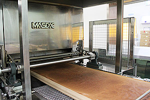 A paper roll sheet where some depth is created using a paper folding machine is laid out. The System Depoly deposits the batter on the sheet, the oven bakes the batter, and then the baked cake is cut both horizontally and vertically. All of these processes are fully automated.