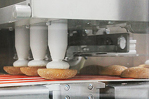 The System Depoly deposits the batter on the paper roll sheet, powdered sugar is applied, the oven bakes the batter, the cream is deposited, and then the cream is sandwiched.